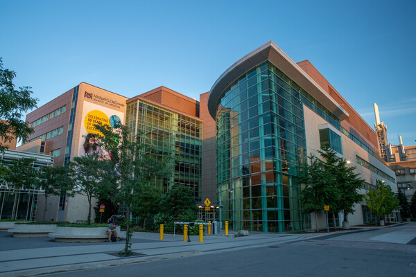 The Michael G. DeGroote Centre for Learning and Discovery (MDCL) building at McMaster University. The building features a modern design with extensive use of glass windows. The surroundings include trees and a pedestrian walkway and on the side of the building, there is a large banner illustrating a sample of MPS's building signs.